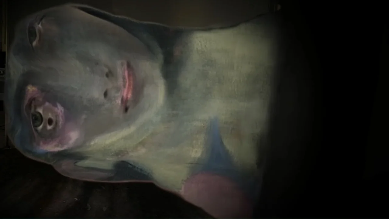 Inland Empire (2006) movie still: closeup — A dark indoor space contains a person with a painted or textured face. The subject rests their head flat…