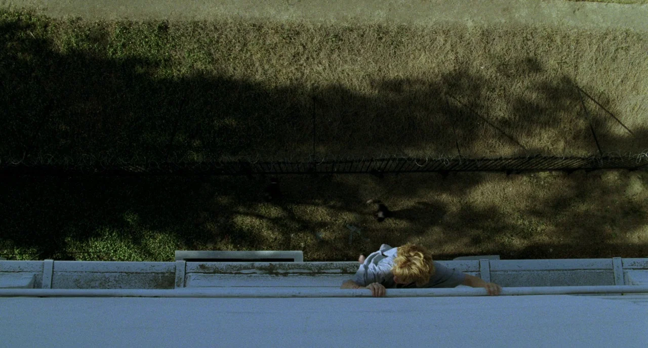 Candyman (1992) movie still: wide — A top-down view shows a person hanging onto a narrow concrete ledge high up. Below lies a strip of dry grass…