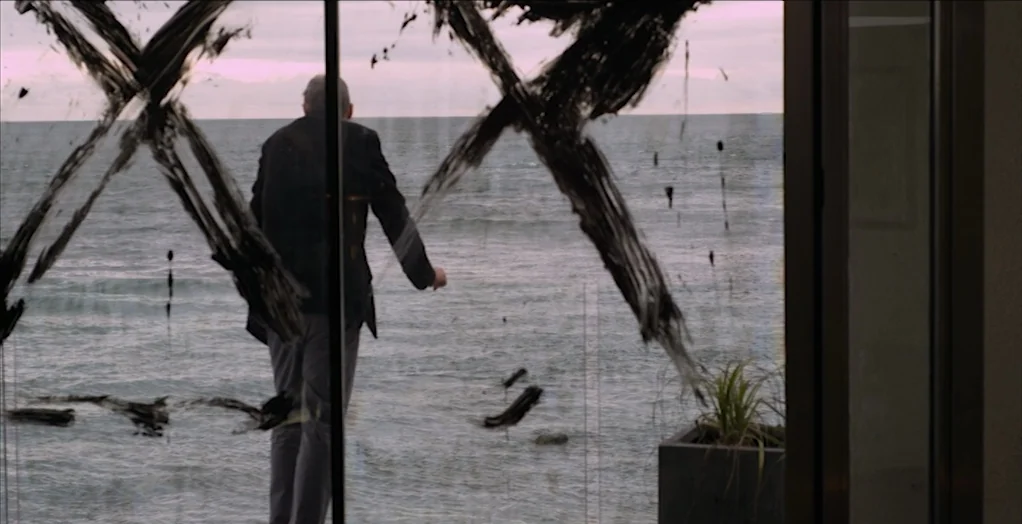 Dollhouse (2012) movie still: fullbody — A modern room features a large glass wall overlooking a calm, grey sea. Thick black paint marks in the…