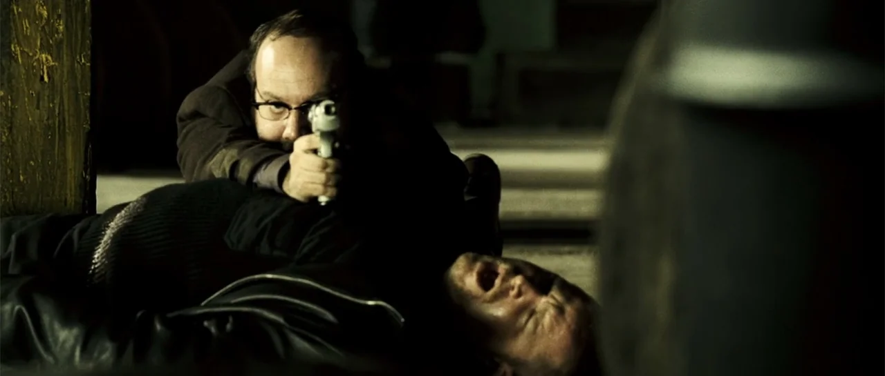 Shoot 'Em Up (2007) movie still: medium — A dimly lit industrial space with dark textures and shadows framing the subjects. One man points a handgun…