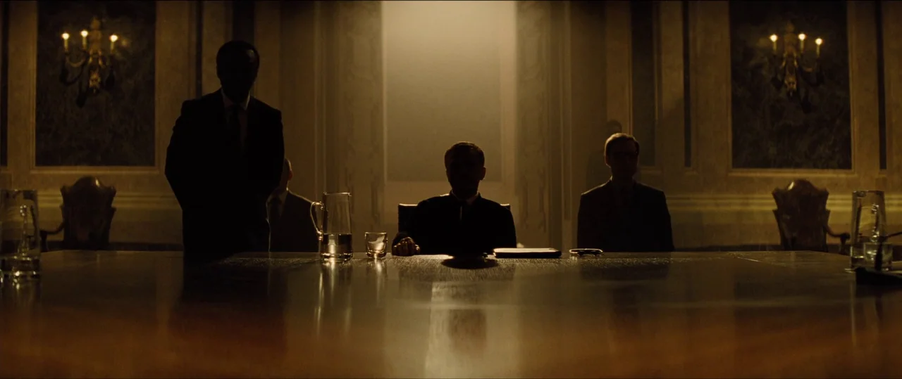 Spectre (2015) movie still: wide — A dark boardroom is lit by a bright doorway and wall-mounted brass sconces. Three men sit as silhouettes around a…