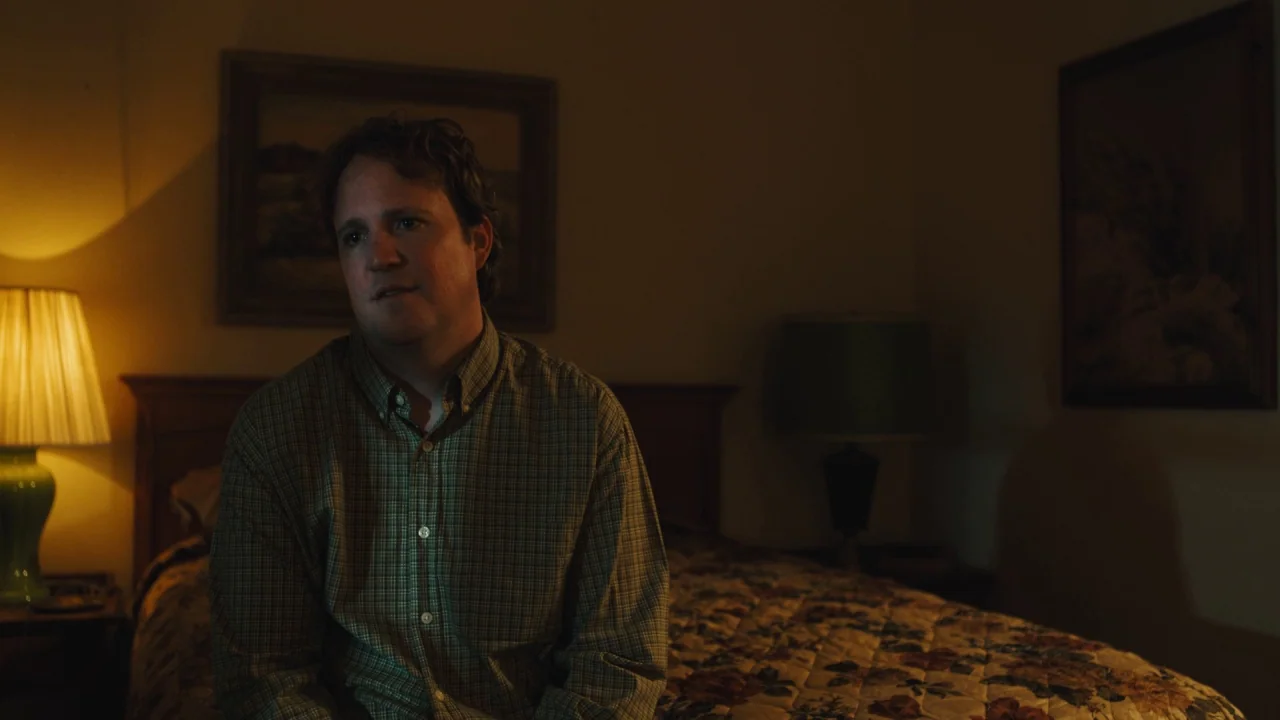 Cam (2018) movie still: medium — A dimly lit room contains a bed with a floral quilt and wall artwork. A man in a plaid shirt sits on the bed edge…