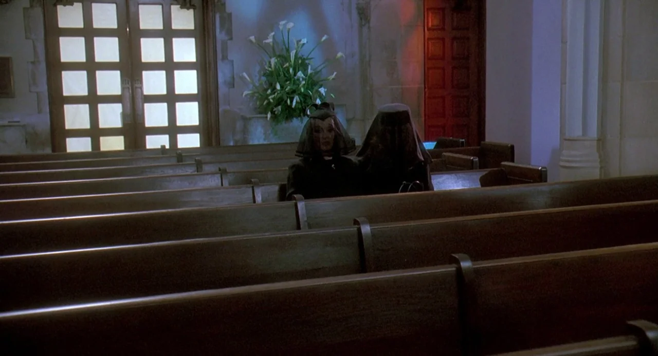 Death Becomes Her (1992) movie still: wide — The interior of a traditional church features rows of wooden pews and arched doorways. Two women in…