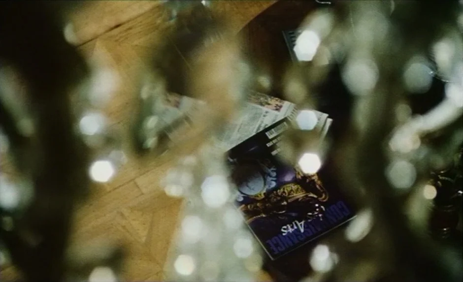 Simon, The Magician (1999) movie still: closeup — A magazine and newspaper lie on a light-colored wooden floor under a chandelier. The view is…