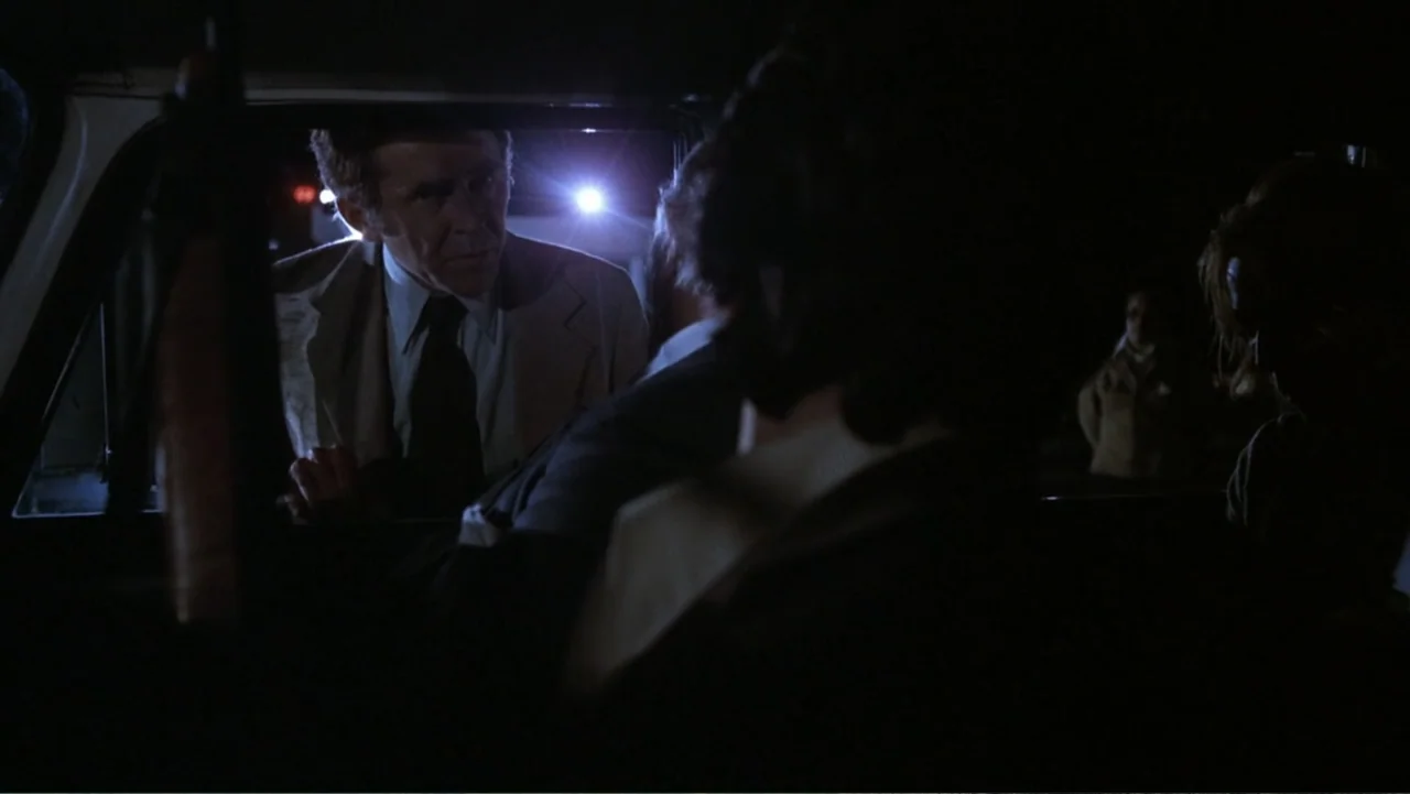 Dog Day Afternoon (1975) movie still: medium — A dark night setting features a person leaning into a car window. An adult male in a light suit…