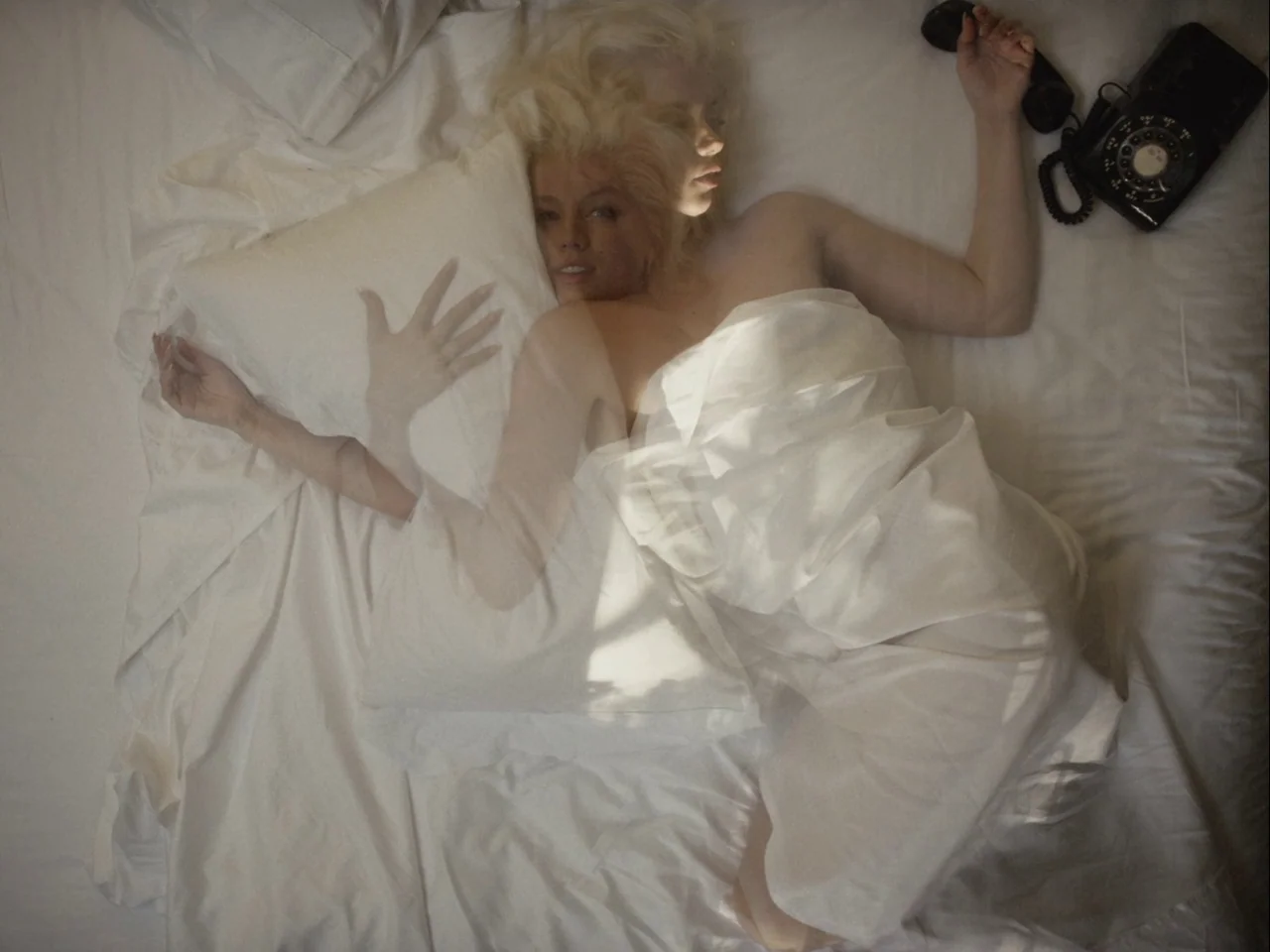 Blonde (2022) movie still: medium — A woman lies on white sheets with a black rotary telephone nearby. Double exposure effects show her face…