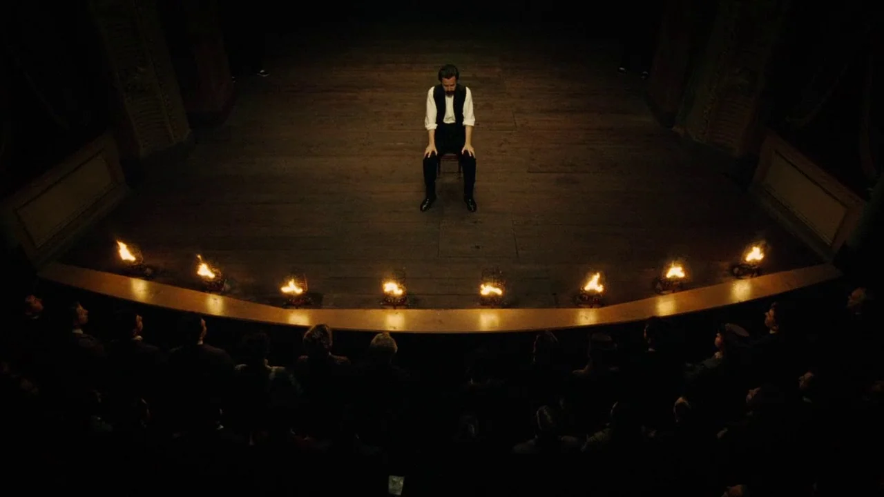 The Illusionist (2006) movie still: wide — A dark, wood-floored stage features a single man seated on a chair. A row of burning fire baskets…