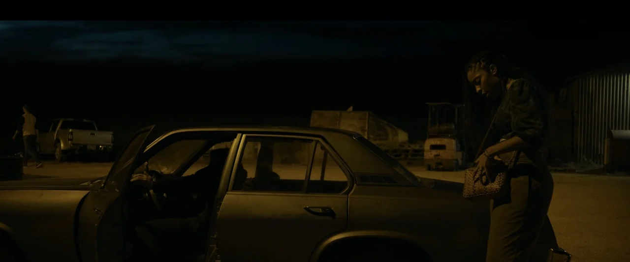 Road House (2024) movie still: wide — An exterior nighttime parking lot features a parked sedan with an open door. A young woman stands at the rear…