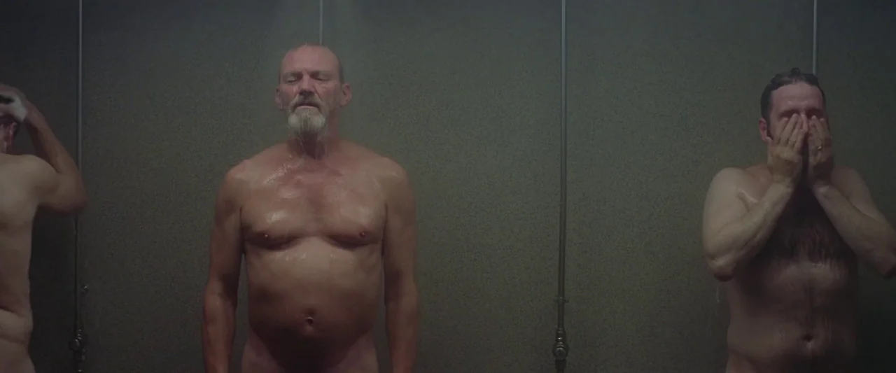 A White, White Day (2019) movie still: medium — A sterile communal shower area features grey tiled walls and vertical plumbing fixtures. Three men…