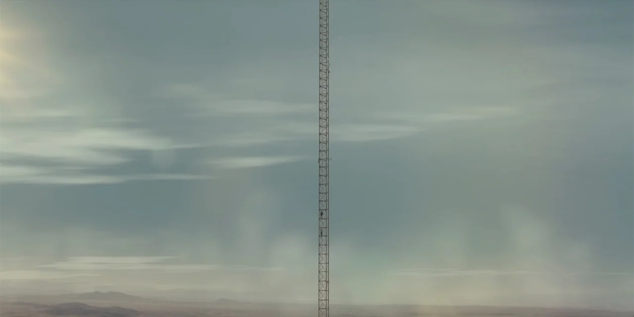 Fall (2022) movie still: establishing — A vast, flat desert landscape stretches under a thick, overcast, grey sky. A solitary, slender, metal…
