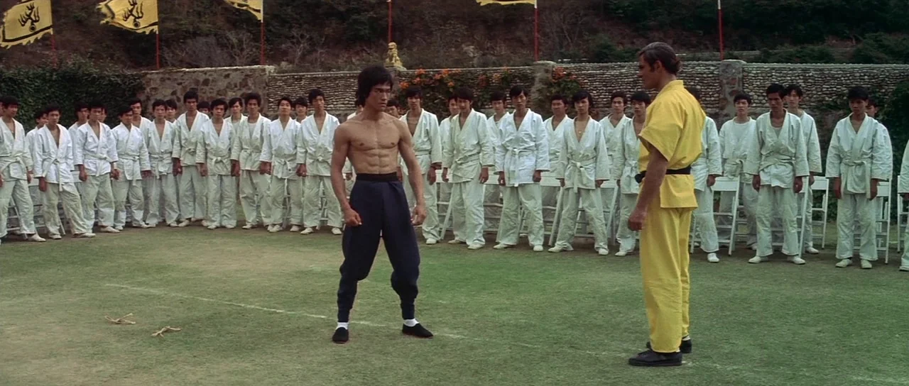 Enter The Dragon (1973) movie still: fullbody — A green training field is surrounded by a low stone wall and yellow flags. Two men in martial arts…