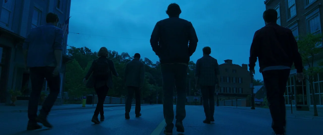 It Chapter Two (2019) movie still: wide — Five figures walk down the middle of a paved bridge during the blue hour. The surroundings include brick…