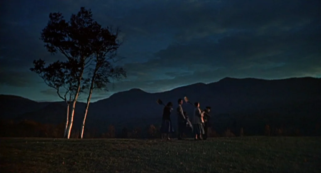 The Trouble With Harry (1955) movie still: wide — A dark grassy meadow sits before a vast range of layered mountains at dusk. Four people walk in a…