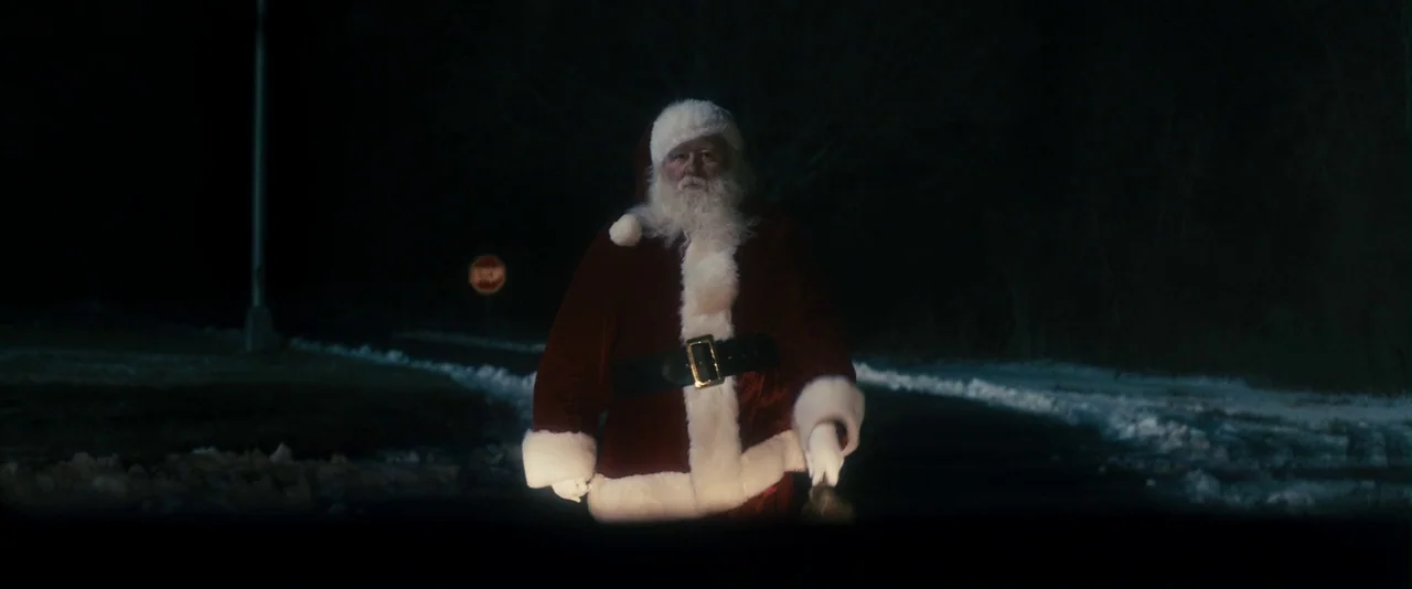 The Box (2009) movie still: fullbody — A man dressed as Santa Claus stands in the middle of a dark road. Snow patches border the dark asphalt under…