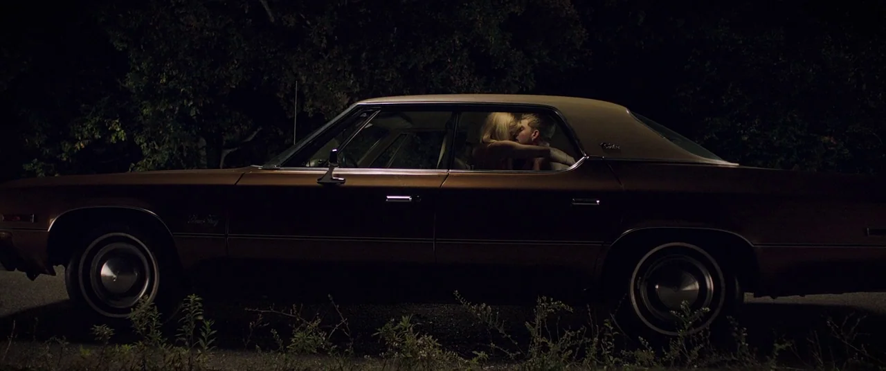 It Follows (2015) movie still: wide — A dark night setting features a brown sedan parked on the side of a road. A couple is visible through the car…
