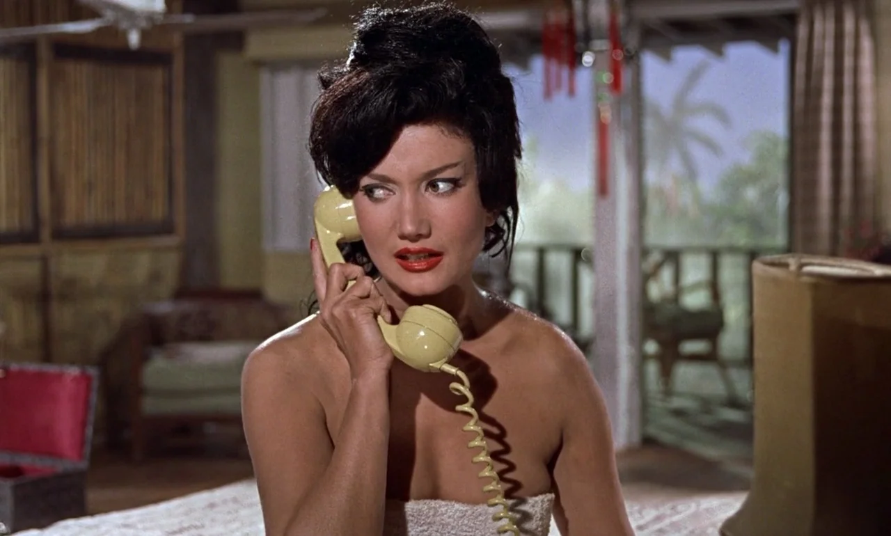 Dr. No (1962) movie still: medium — A 1960s hotel room features bamboo furniture and an open balcony door overlooking trees. A woman with a dark…