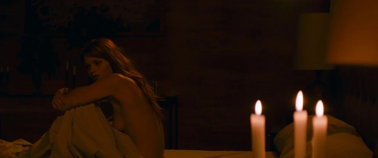 Elizabeth Harvest movie still: medium — A dark, dimly lit bedroom at night features warm candlelight illuminating the subject. A woman sits in bed…