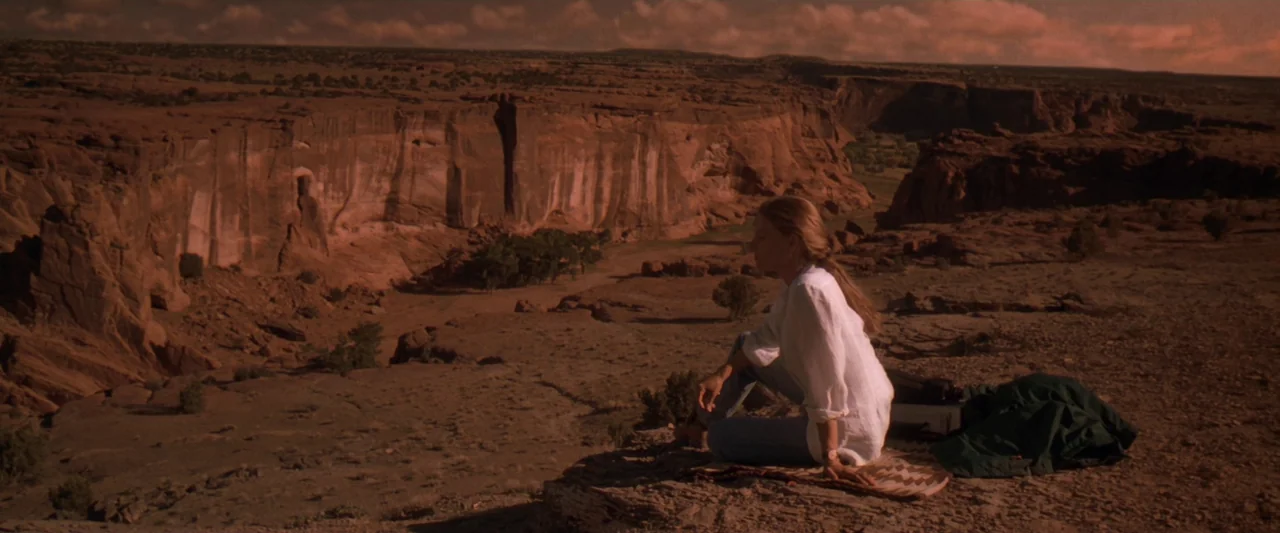 Contact (1997) movie still: wide — A vast, arid desert canyon with deep sandstone cliffs and scrub vegetation. A woman sits on a rock ledge while…