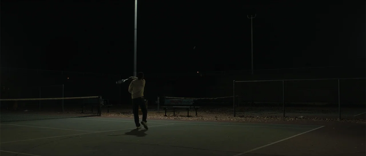 King Richard (2021) movie still: wide — A dark outdoor tennis court at night is lit by a single tall streetlamp. A solitary man in a light sweater…