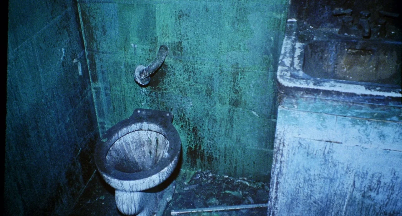 Candyman (1992) movie still: wide — A grimy, neglected bathroom features weathered teal tiling and a stained porcelain toilet. Stains and debris…