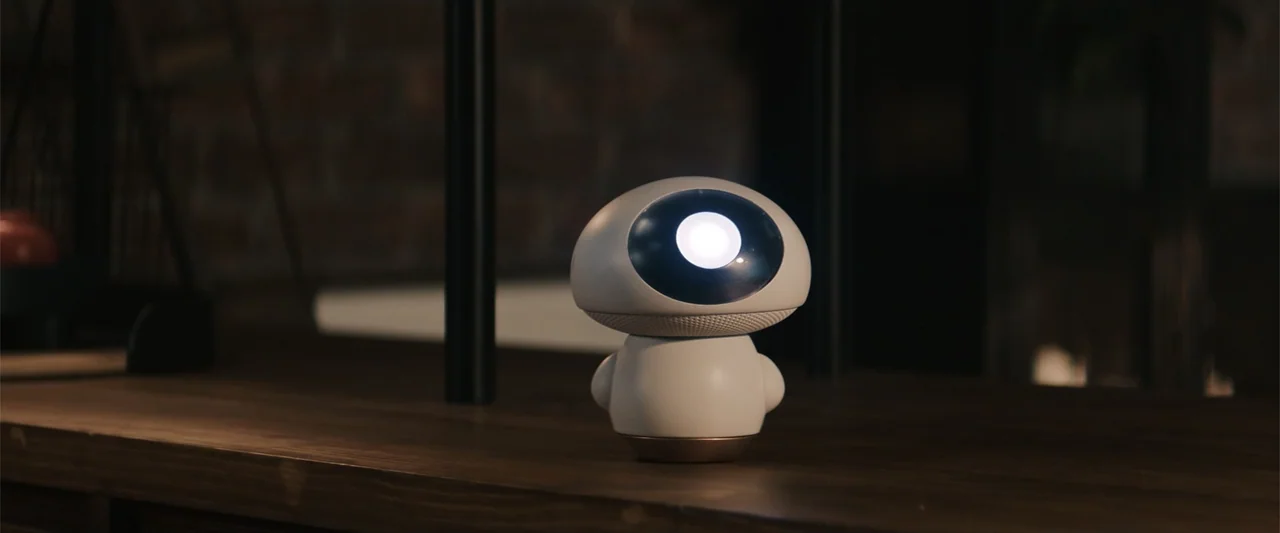 M3GAN 2.0 (2025) movie still: medium — A small white robot with a glowing circular face sits on a dark wooden surface. The background features…