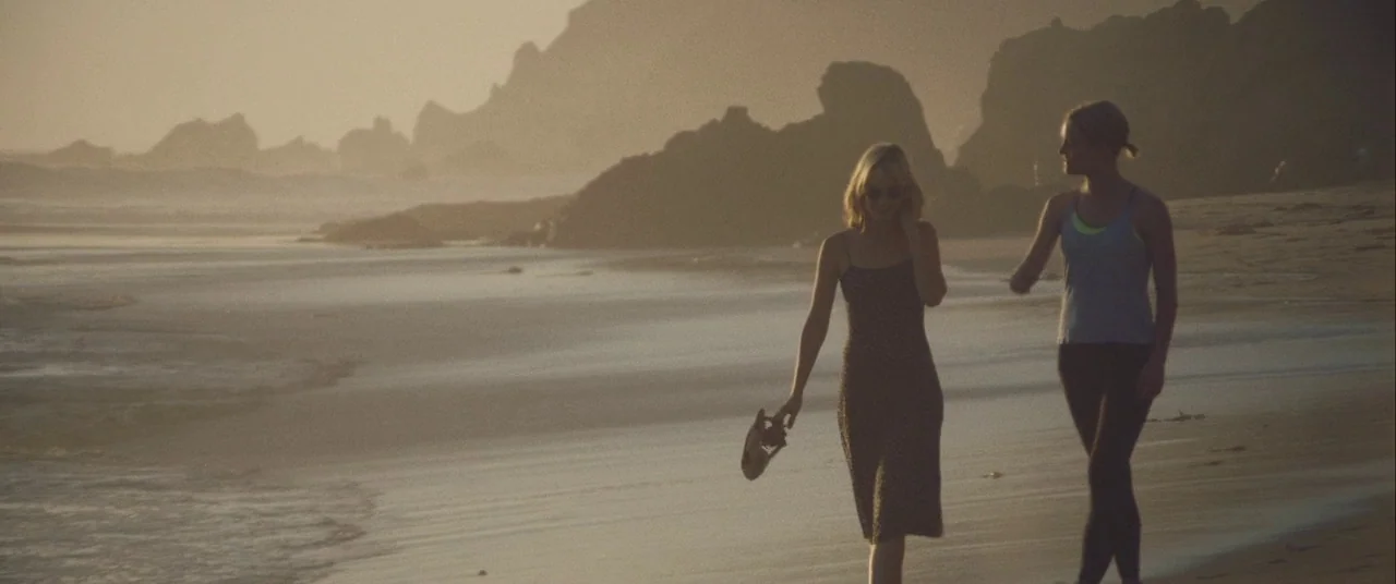 Always Shine (2016) movie still: fullbody — A wide beach at sunset features large rock formations in the hazy distance. Two young women walk…