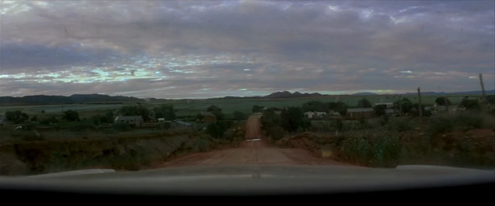 Silent Light (2007) movie still: establishing — A rural landscape unfolds under an overcast sky with a dirt road leading ahead. Sparse vegetation…