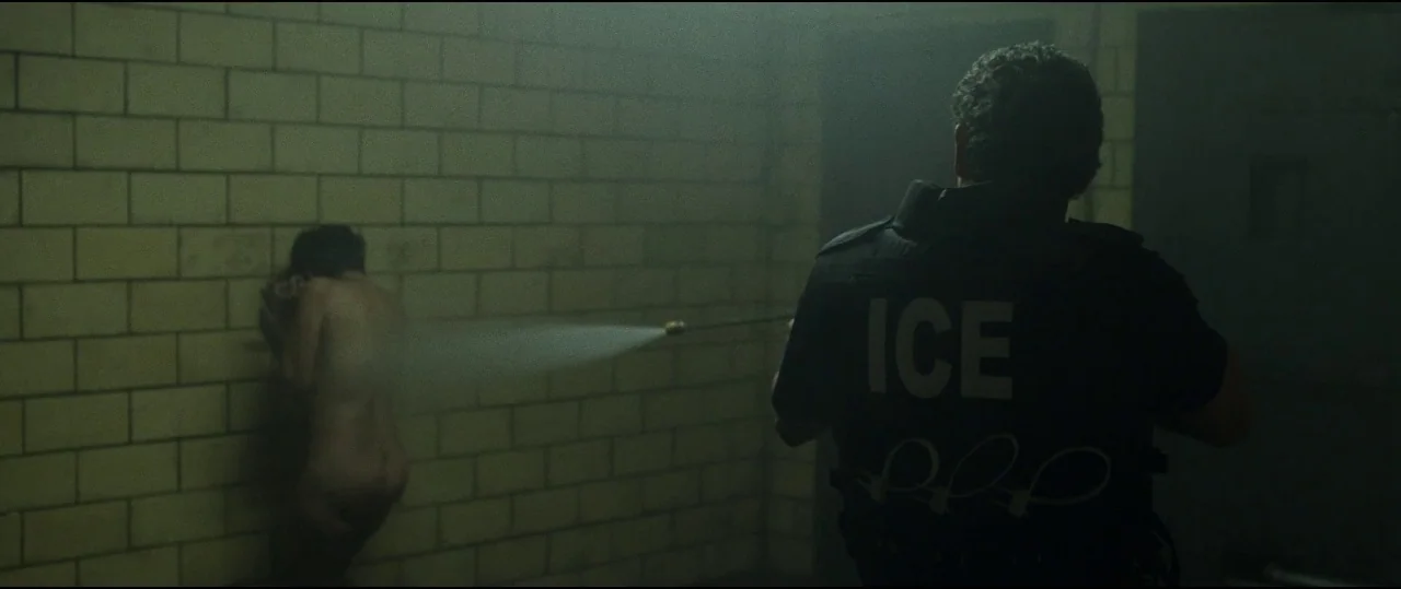 Lowlife (2017) movie still: medium — A dimly lit room with yellow brick walls and heavy, shadowed corners. An officer in an ICE vest directs a…