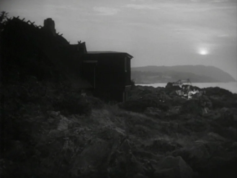 A Lesson In Love (1954) movie still: establishing — A dark wooden structure sits on a rugged, uneven hillside overlooking the ocean water. The…
