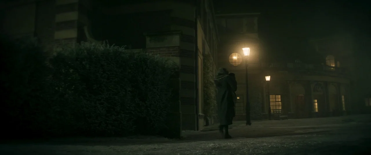 Wonka (2023) movie still: wide — A dark, foggy city street at night features red brick buildings and a large hedge. A lone figure in a long coat…