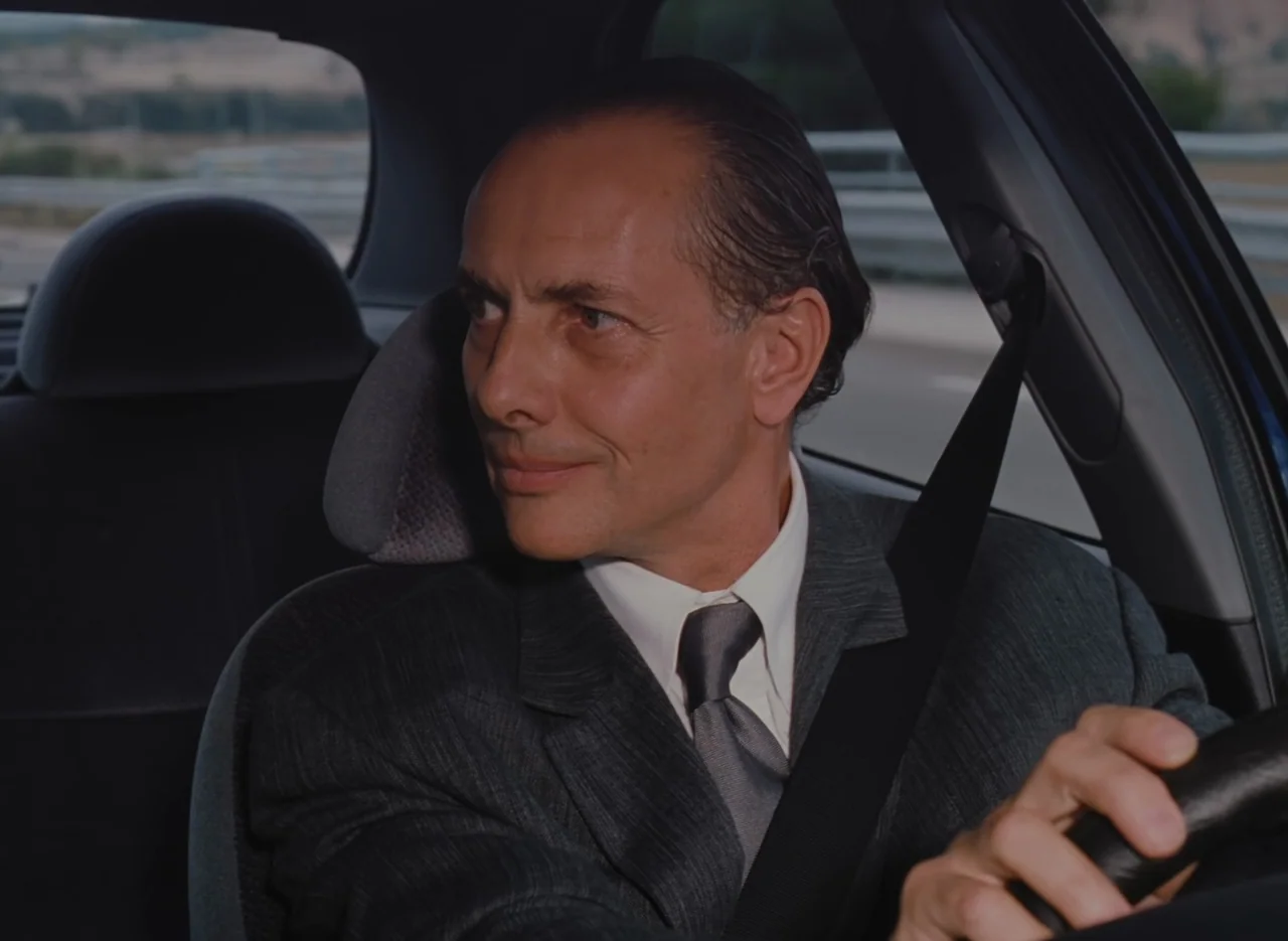 Autumn Tale (1998) movie still: medium — A man drives a vehicle on a road with metallic guardrails visible outside. The driver wears a formal suit…