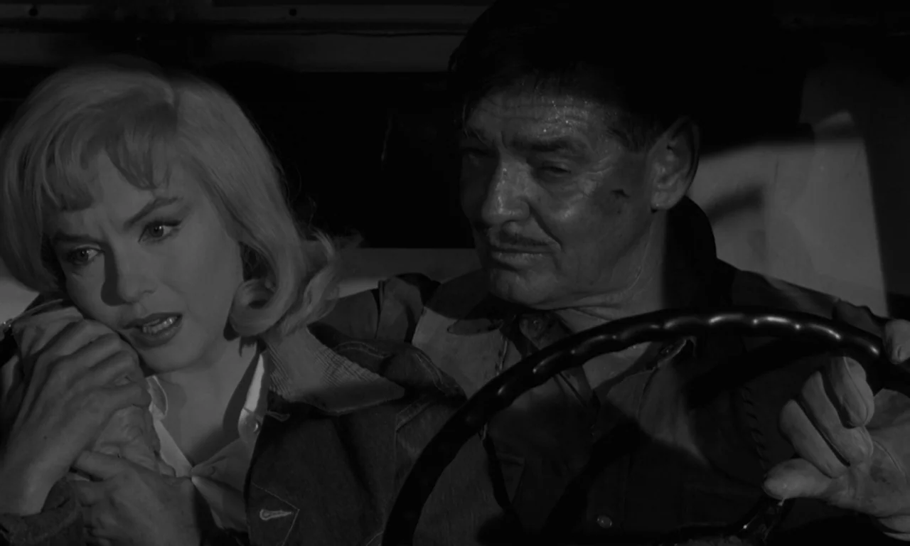 The Misfits (1961) movie still: medium — Two people sit inside the darkened cab of a vehicle at night. The man keeps his hands on the steering wheel…
