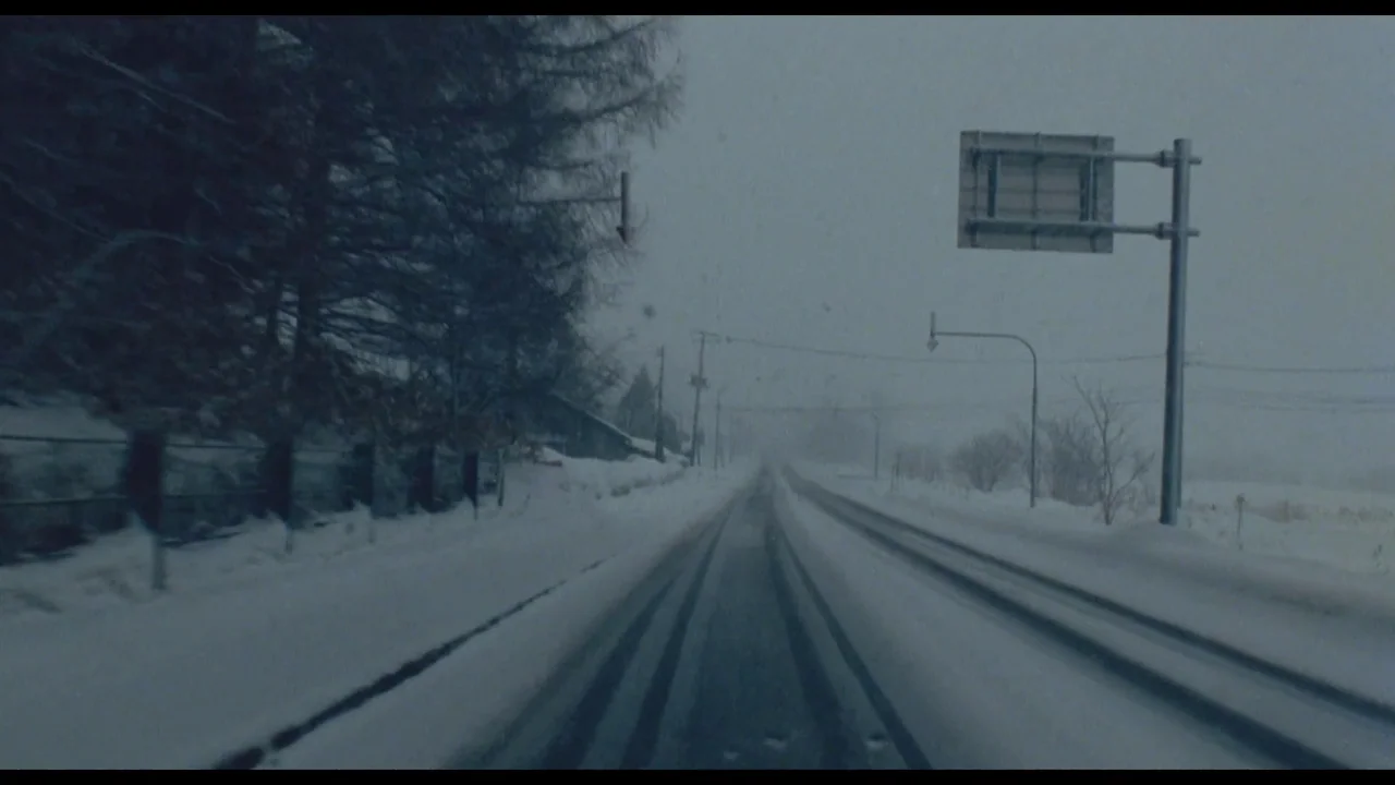 Millennium Mambo (2001) movie still: wide — A desolate two-lane road disappears into heavy snow and fog during the daytime. Dark tire tracks cut…