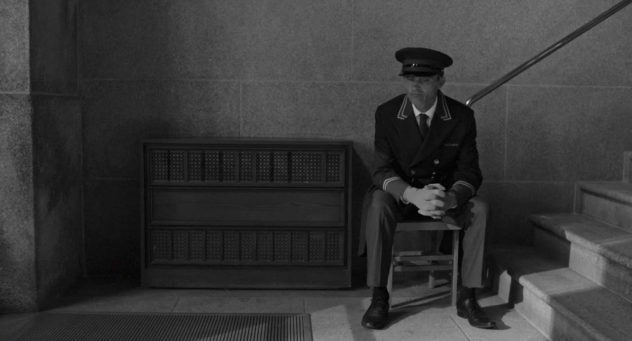 Song of Granite (2017) movie still: medium — A stone stairwell features a heavy wooden radiator cover mounted against a concrete wall. A uniformed…