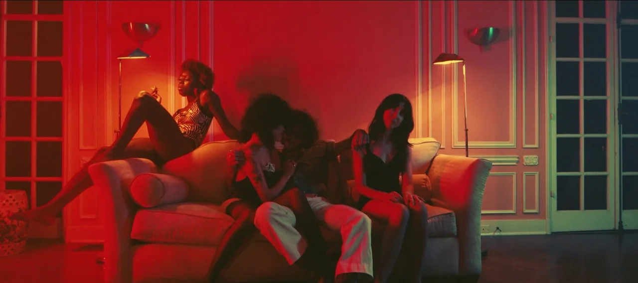 Fuzzy Head (2023) movie still: wide — An opulent living room with white molding is bathed in deep monochromatic red light. Four people lounge across…