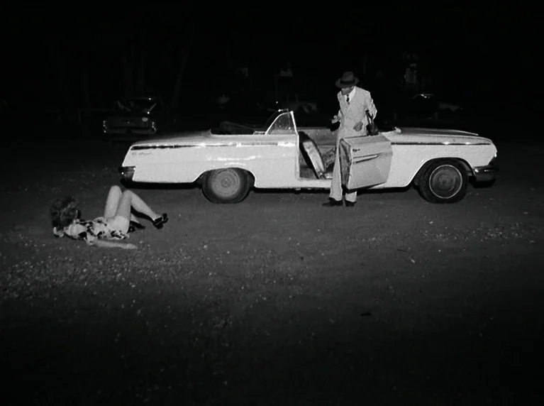 The American Soldier (1970) movie still: wide — A dark, gravel-covered parking lot at night features a white vintage convertible car. A man in a…