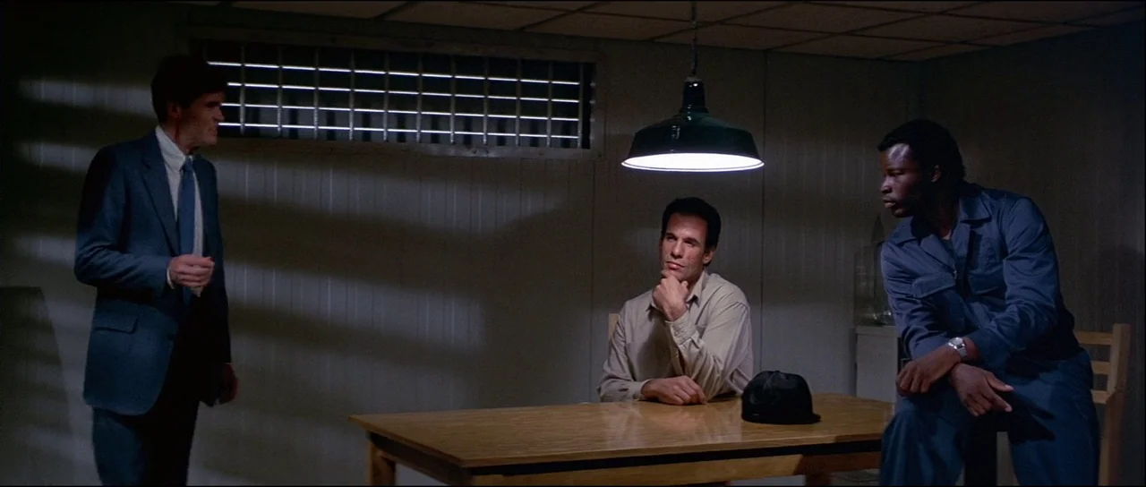 Licence to Kill (1989) movie still: wide — A spartan room features wood-paneled walls and a high-set grated window. Three men interact around a…