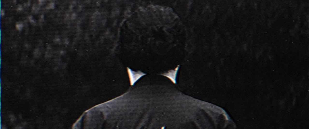 Broadcast Signal Intrusion (2021) movie still: medium — A dark environment surrounds a person seen from the back in silhouette. The frame features…