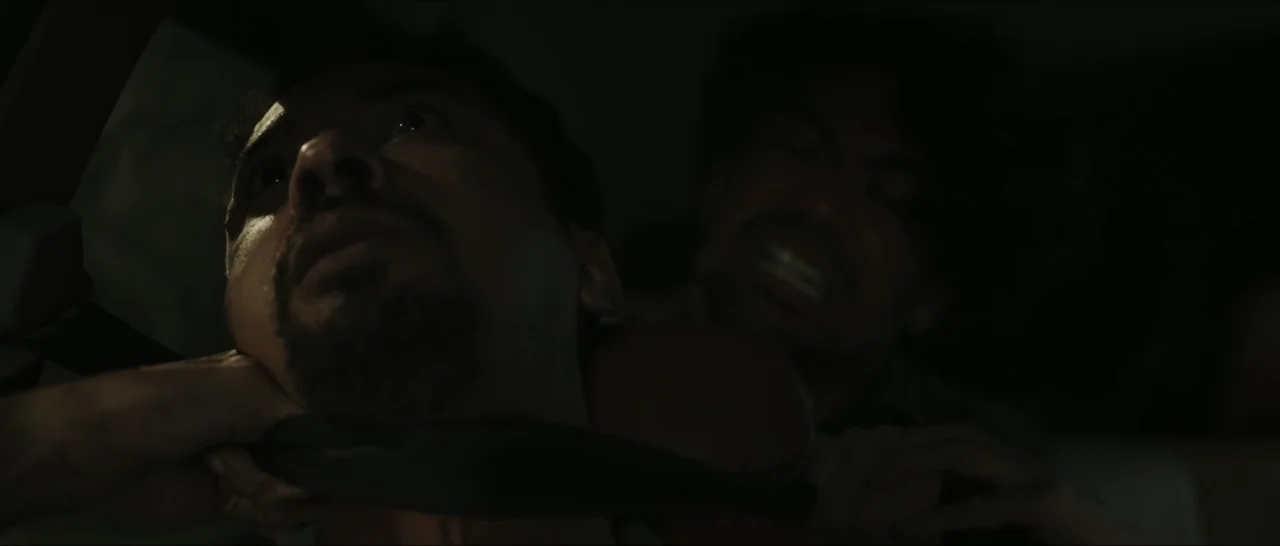Innocent Vengeance (2023) movie still: closeup — The interior of a dark vehicle at night with very limited visibility. One man pulls a strap tightly…
