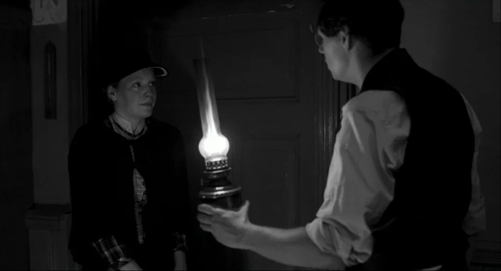 The White Ribbon (2009) movie still: medium — A dimly lit indoor hallway with a wooden paneled wall and a door frame. A man in a vest holds a…