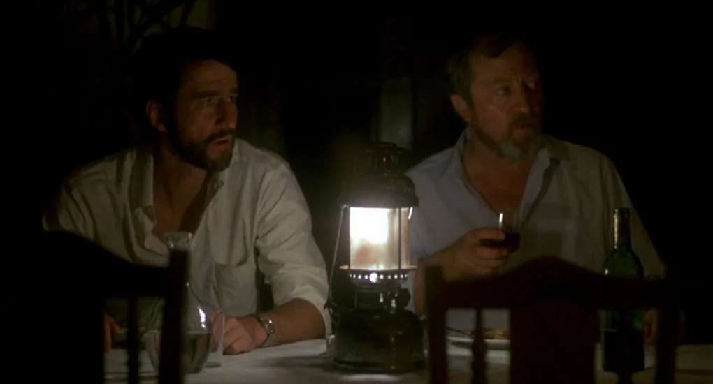 The Killing Fields (1984) movie still: medium — Two men sit across from each other at a table in a dark room. An oil lantern between them…