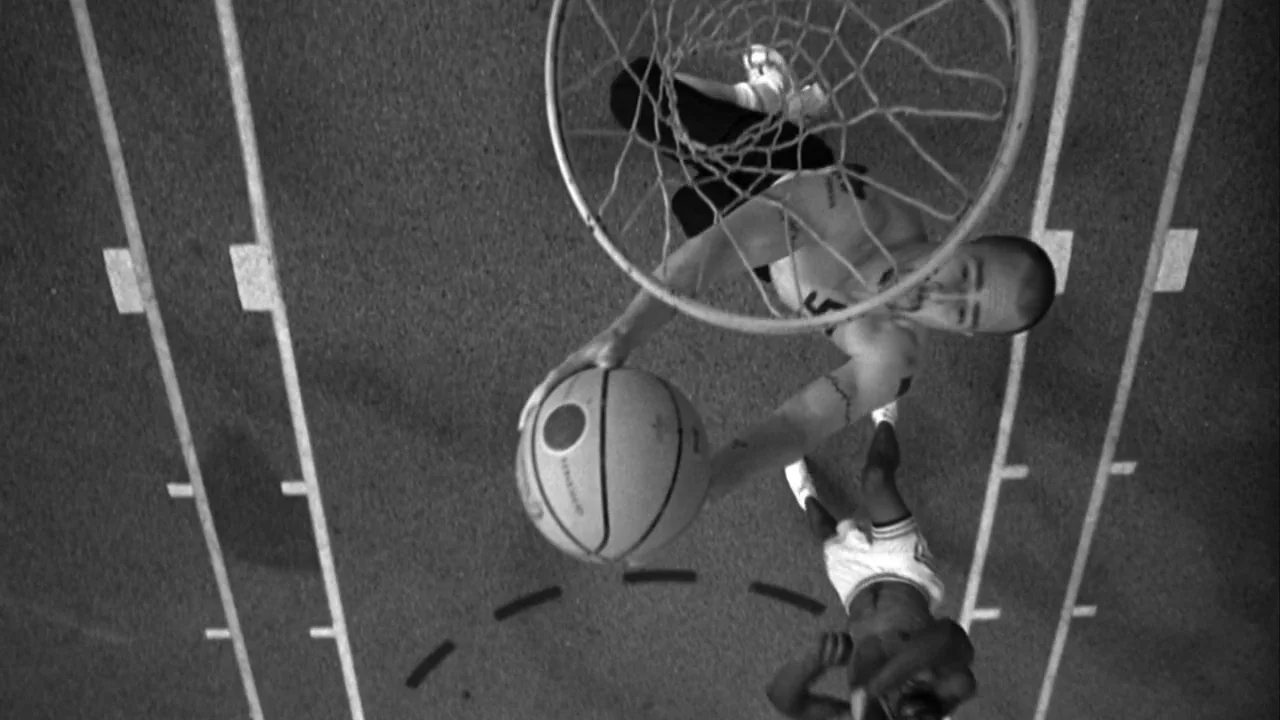 American History X movie still: wide — An outdoor court with marked white lines on a dark asphalt surface at night. A player dunks a basketball…