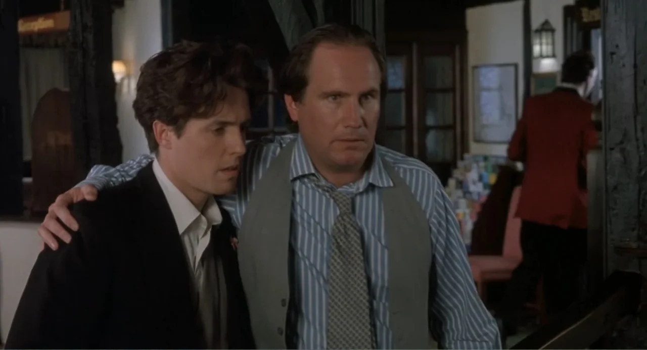 Four Weddings and a Funeral (1994) movie still: medium — A dimly lit hotel lobby features dark wooden structural beams and a background desk. A man…