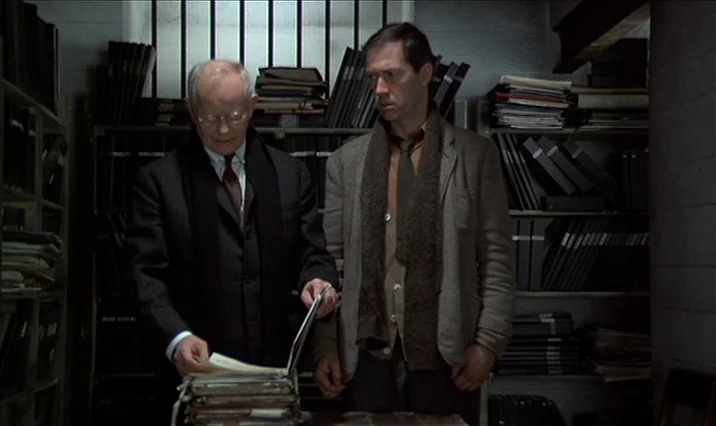 The Serpent’s Egg (1977) movie still: medium — A dimly lit room is filled with rows of metal shelving holding numerous binders. Two men stand at a…
