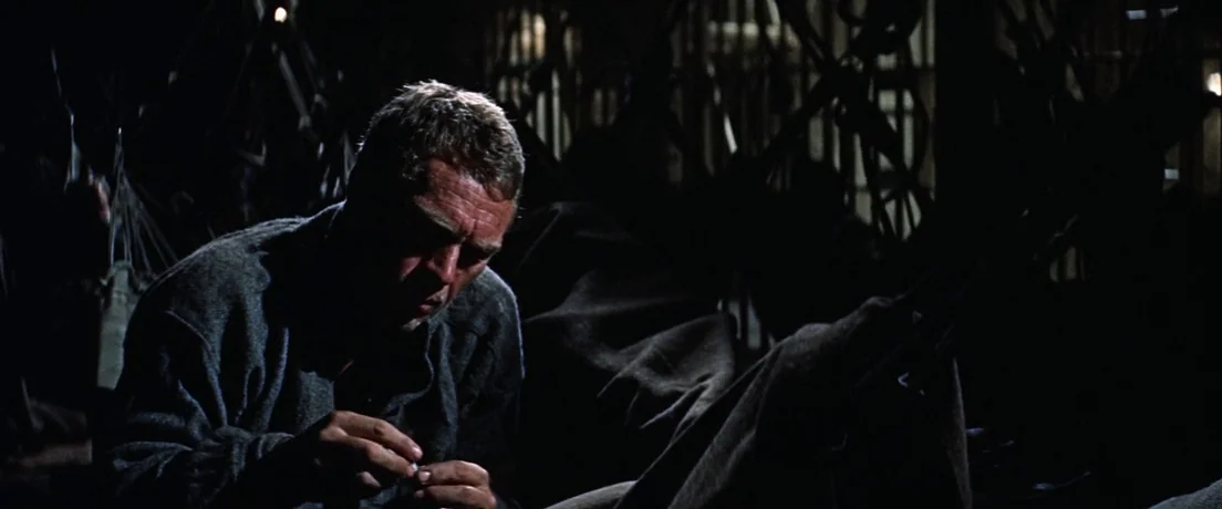Papillon (1973) movie still: medium — A dimly lit, sparse bunkhouse room with a dark wooden structure visible in the background. A man sits alone on…