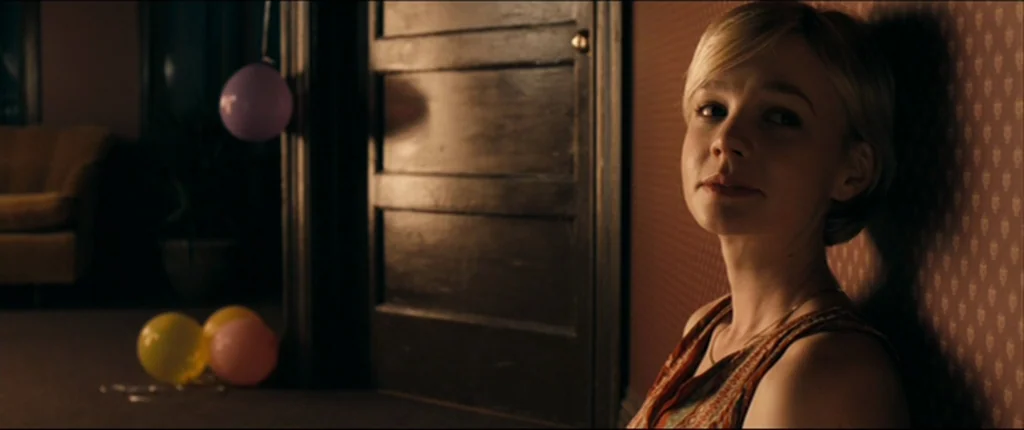 Drive (2011) movie still: medium — A dimly lit residential space with patterned wallpaper features a closed dark wooden door. A young woman with…