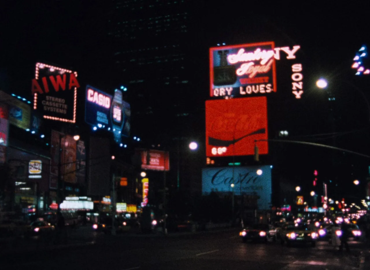 Basket Case (1982) movie still: establishing — A dark urban intersection at night is filled with brightly lit, vintage neon advertising signs. Cars…