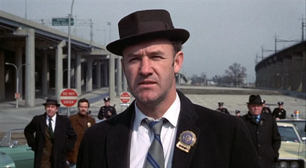 The French Connection (1971) movie still: medium — A man in a fedora stands in front of a sprawling elevated concrete highway system. Several other…