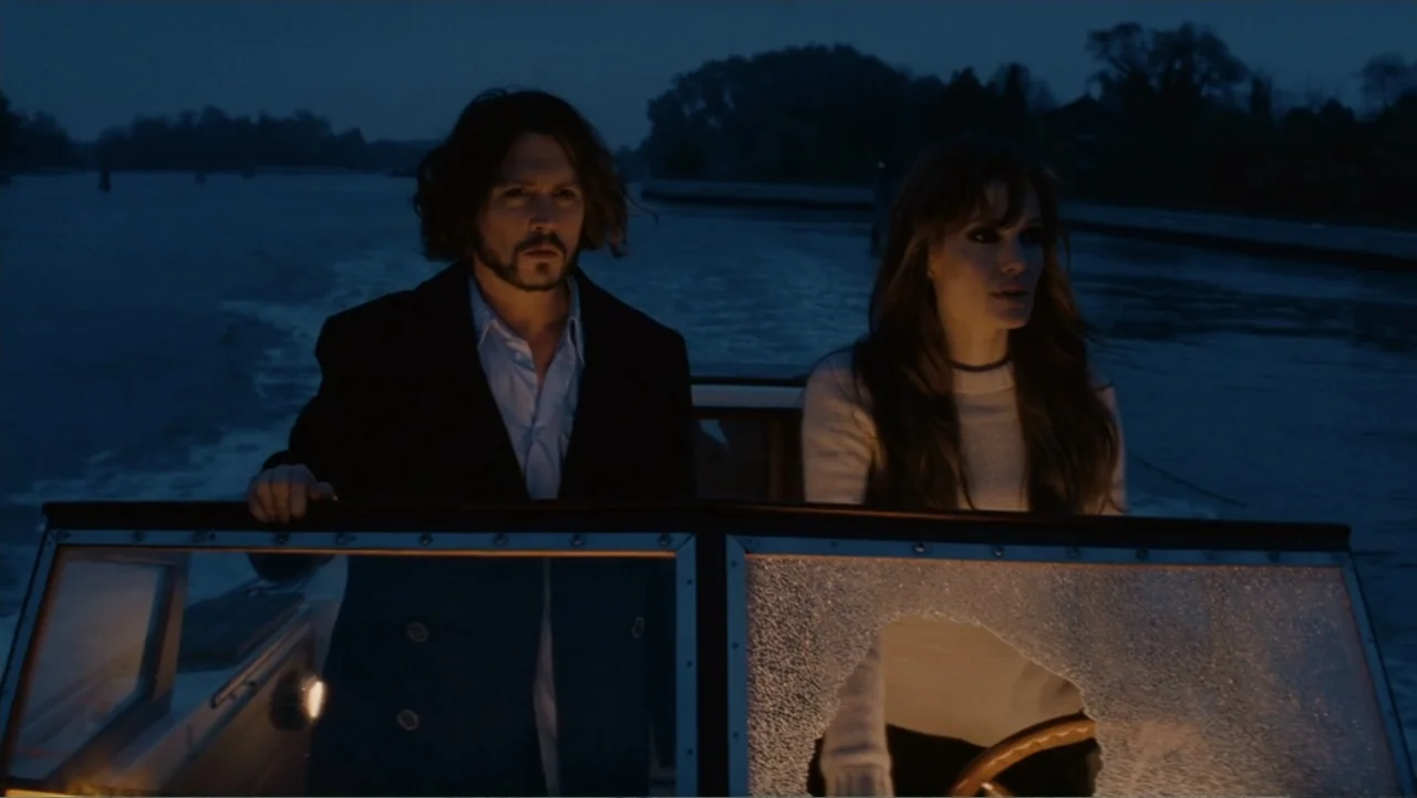The Tourist (2010) movie still: medium — Two people travel across dark water on a motorboat during the late evening hours. They stand behind the…