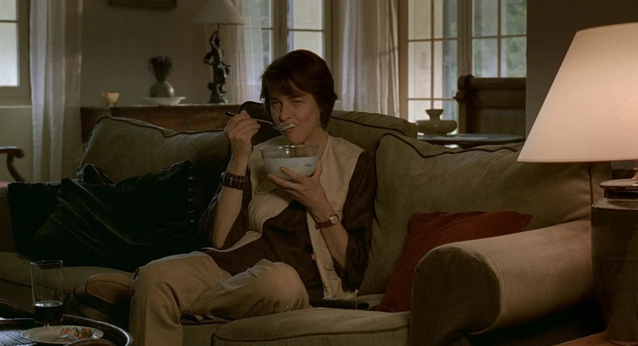 Swimming Pool (2003) movie still: medium — A dimly lit room with a beige sofa and wooden side furniture. A woman sits on the sofa and eats food from…